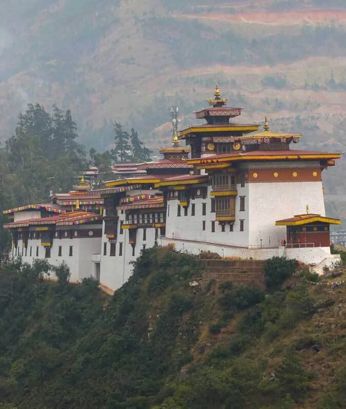 Un long bâtiment blanc composés de nombreux bâtiments dans un style traditionnel bhoutanais, longeant la crête de la montagne. Les toits sont dans le même style, en rouge et jaune.