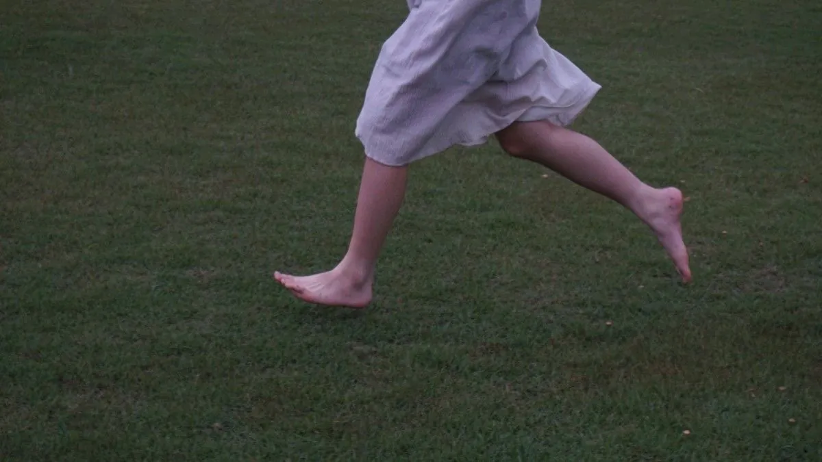 Sur de l’herbe bien taillée, court une silhouette dont on ne voit que le bas des jambes et la robe légère blanche