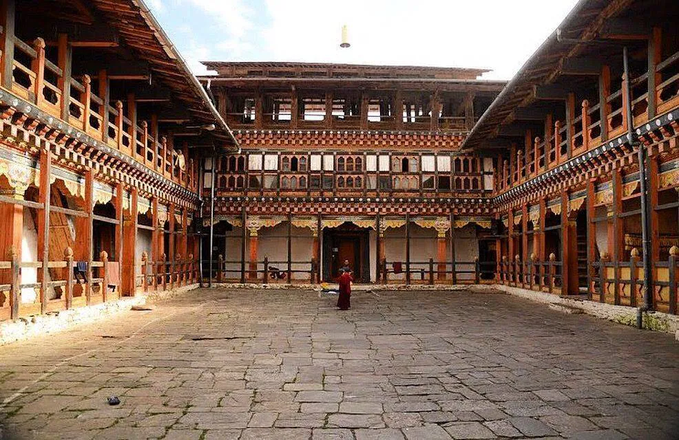 Une cour ouverte pavée de pierres, entourée par trois façades montrant des couloirs extérieurs composées de bois rouge et jaune. Des décorations discrètes sont présentes.